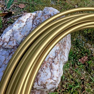 May include: Close-up of several gold-colored metal hoops, possibly for crafting or decoration. The hoops are arranged in a curved formation, with a smooth, reflective surface. The background features a large, textured rock and patches of green grass.