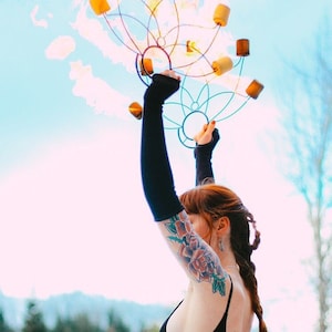May include: A woman with red hair and a floral tattoo on her arm is performing with a fire spinning apparatus. The apparatus is made of metal and has multiple points where flames can be attached. The woman is wearing black gloves and a black top.