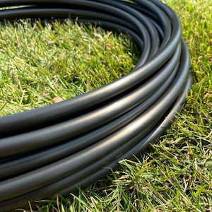 May include: Coiled black tubing, likely for irrigation or plumbing, rests on a bed of green grass. The tubing is smooth and appears to be made of a flexible material. The image is taken outdoors in natural light.