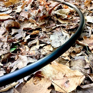 May include: A smooth, black, circular tubing segment made of flexible material, curved in a wide arc. The surface of the tube is solid black with a slight sheen, showing a consistent cylindrical shape throughout the visible length of the object.