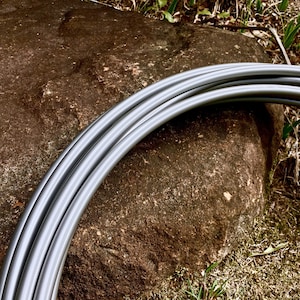 May include: Curved, gray metal tubing, possibly for a craft or construction project, is arranged in a semi-circle. The tubes are smooth and reflective, with a metallic sheen. The background features a large, textured brown rock and patches of green grass.