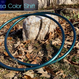 May include: Two teal-colored metal hoops, also known as hula hoops, are lying on the ground. The hoops are partially overlapping and are resting on a bed of brown leaves and green grass. The text "Medusa Color Shift" is visible in the background.
