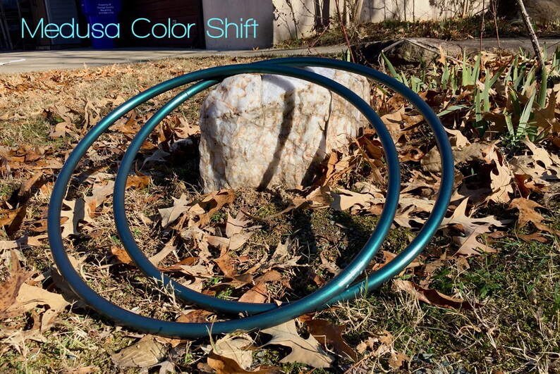 May include: Two teal-colored metal hoops, also known as hula hoops, are lying on the ground. The hoops are partially overlapping and are resting on a bed of brown leaves and green grass. The text "Medusa Color Shift" is visible in the background.