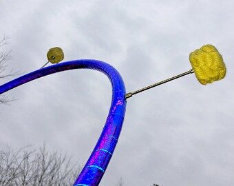 May include: A blue iridescent hoop with two yellow yarn pom-poms attached to the hoop with metal rods.