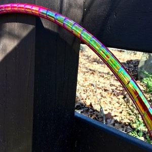May include: A close-up of a pink and iridescent hula hoop with a segmented design. The hoop is partially visible against a dark wooden background.