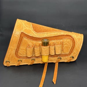 May include: A tan leather stock cover featuring a textured finish, designed with five integrated loops for ammunition storage. One yellow shotgun shell is inserted into the center loop. The cover includes brass eyelets and leather lacing along the bottom edge.