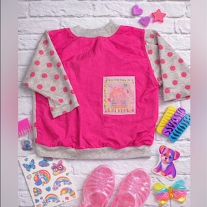 May include: A bright pink long-sleeve shirt with light grey sleeves featuring pink polka dots. The shirt has a light grey ribbed collar and hem, and a rectangular patch on the front displaying a cartoon character and the text "BEAR".