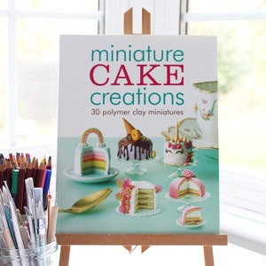 Puede incluir: Un libro titulado "miniature CAKE creations" con un fondo azul verdoso y un título blanco. La portada del libro presenta 30 miniaturas de pasteles de arcilla polimérica, incluyendo un pastel de arcoíris, un pastel de unicornio y un pastel de chocolate con un cono en la parte superior.
