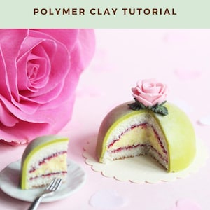 Puede incluir: Un tutorial de arcilla polimérica para hacer un pastel de princesa sueca en miniatura. El pastel es verde con una rosa rosa en la parte superior y está cortado por la mitad para mostrar las capas de crema amarilla y mermelada de frambuesa. El pastel está sobre un encaje blanco con corazones rosas esparcidos a su alrededor.