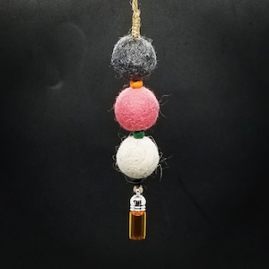 Op de afbeelding: Een hangende aromatherapie-diffuser met drie vilten ballen in grijs, roze en wit, geregen aan een natuurlijke vezelkoord. Een kleine glazen fles met een zilveren dop en amberkleurige vloeistof hangt aan de onderkant.