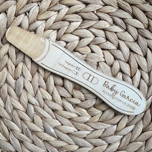May include: A light-colored wooden pregnancy test replica featuring engraved text that reads Pregnant, Not Pregnant, Baby Garcia, and Arriving March 2026, with two vertical lines indicating a positive result on the display area.