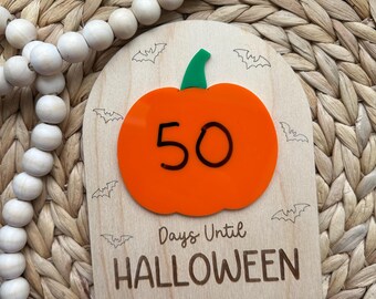 May include: Wooden Halloween countdown sign featuring an orange pumpkin with the number 50 and the text "Days Until Halloween." The sign has a green stem and bat silhouettes. The sign is on a woven background.