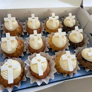 May include: A box of cupcakes with white frosting and white cross decorations. The cupcakes are in decorative paper cups and have a variety of cake colors, including brown, tan, and reddish-brown. The box is white with a blue interior.
