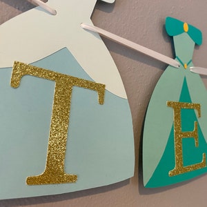 May include: A princess-themed banner with the letters "T E L" in gold glitter on a blue, green, and yellow background. The letters are attached to a white ribbon.