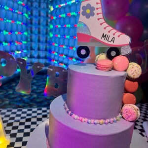 May include: A two-tier lavender cake decorated with a string of small colorful beads and a vertical stack of assorted macarons. A white roller skate cake topper with a blue flower and the name MILA is placed on top.