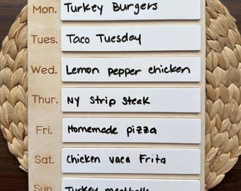May include: A wooden weekly meal planner with white removable labels. The labels are written with a black marker and list the meals for each day of the week. The days of the week are written in brown on the wood. The meals are: Monday - Turkey Burgers, Tuesday - Taco Tuesday, Wednesday - Lemon Pepper Chicken, Thursday - NY Strip Steak, Friday - Homemade Pizza, Saturday - Chicken vaca Frita, Sunday - Turkey Meatballs.