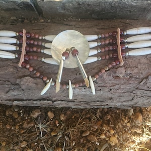May include: A multi-strand necklace with white bone beads, brown leather, and red glass beads. The necklace has a large, round, white shell pendant with a silver design.