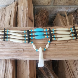 May include: A handcrafted choker necklace featuring off-white bone beads, turquoise accents, and brass-colored spacers. The necklace has a black backing and a pendant. The design is reminiscent of Native American jewelry.