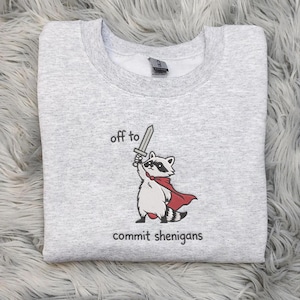 May include: A pale grey sweatshirt featuring a cartoon raccoon with a sword and red cape, with the text "off to commit shenanigans". The design is printed on the front of the garment.