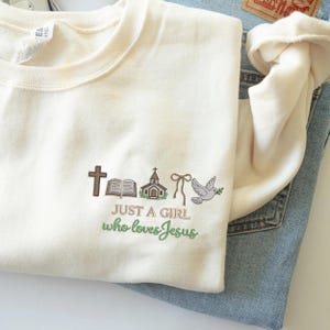 May include: Cream-coloured sweatshirt featuring embroidered religious symbols: a cross, an open book, a church, a bow, and a dove with an olive branch. The text "JUST A GIRL who loves Jesus" is embroidered below.