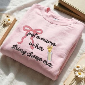 May include: A light pink sweatshirt with the embroidered phrase "just a mama in her string cheese era." The design includes a pink bow and a string cheese graphic. The sweatshirt is folded on a white surface.