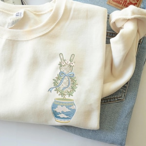 Könnte beinhalten: Cremefarbenes Sweatshirt mit einem gestickten Design eines hasenförmigen Topiaries in einer blau-goldenen Vase. Der Hase hat einen Blumenkranz und ein blaues Band. Das Sweatshirt ist auf einer hellblauen Jeans gefaltet.