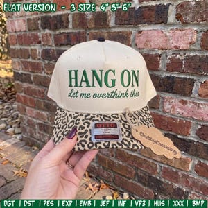 May include: A cream-colored baseball cap with a leopard print brim. The front features the embroidered green text HANG ON and Let me overthink this. An Otto brand sticker is on the brim, and a brown tag labeled ChubbyCheeksDigi is attached.