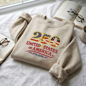 Puede incluir: Una sudadera beige con el texto "250 UNITED STATES OF AMERICA 1776-2026" bordado en rojo, blanco y azul. El diseño presenta el número 250 con un patrón de la bandera estadounidense. La sudadera está doblada sobre una superficie blanca.