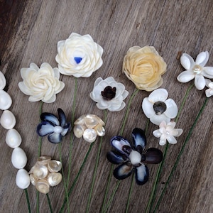 K&ouml;nnte beinhalten: Eine Sammlung handgefertigter Muschelblumen in verschiedenen Formen und Farben, darunter Wei&szlig;, Creme und Dunkelblau. Die Blumen sind mit gr&uuml;nen Stielen auf einer Holzoberfl&auml;che angeordnet.