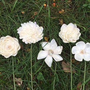 K&ouml;nnte beinhalten: F&uuml;nf elfenbeinfarbene Kunstblumen mit gr&uuml;nen Stielen sind in Gras angeordnet. Die Blumen haben verschiedene Formen, darunter eine Rose, eine Chrysantheme und eine Magnolie. Jede Blume hat eine perlenartige Mitte.