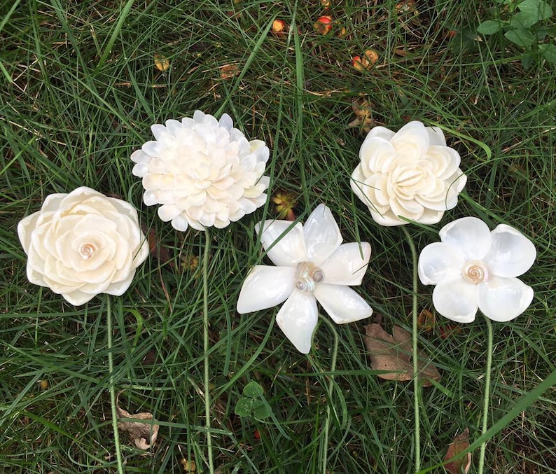 K&ouml;nnte beinhalten: F&uuml;nf elfenbeinfarbene Kunstblumen mit gr&uuml;nen Stielen sind in Gras angeordnet. Die Blumen haben verschiedene Formen, darunter eine Rose, eine Chrysantheme und eine Magnolie. Jede Blume hat eine perlenartige Mitte.