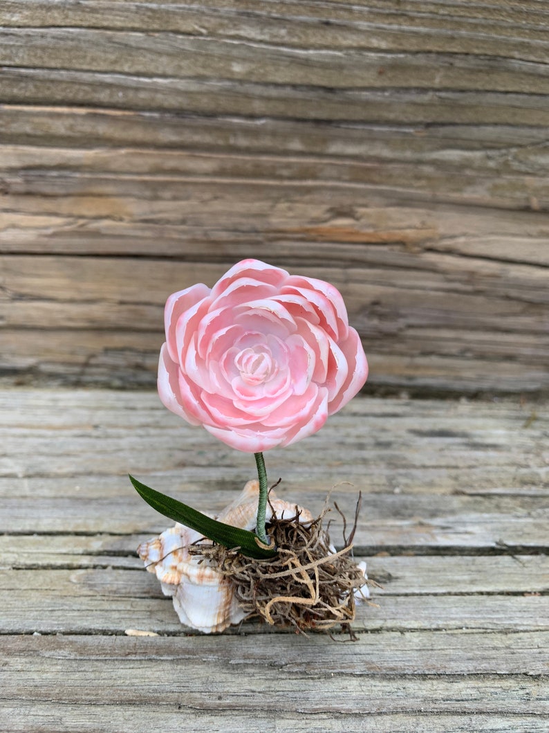 May include: A delicate pink rose made of porcelain sits in a seashell with brown roots and green leaves.
