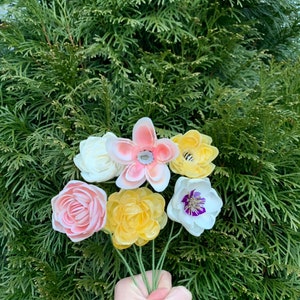 K&ouml;nnte beinhalten: Ein Strau&szlig; bunter Kunstblumen, darunter rosa, gelbe und wei&szlig;e Bl&uuml;ten, vor einem Hintergrund aus gr&uuml;nem Laub. Die Blumen haben detaillierte Bl&uuml;tenbl&auml;tter und sind auf gr&uuml;nen Drahtstielen angeordnet.
