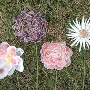 K&ouml;nnte beinhalten: Vier handgefertigte dekorative Blumen aus Muscheln, angeordnet auf Gras. Die Blumen sind in den Farbt&ouml;nen Rosa, Wei&szlig; und Lila gehalten und haben gr&uuml;ne Stiele. Eine Blume hat eine spiralf&ouml;rmige Mitte.