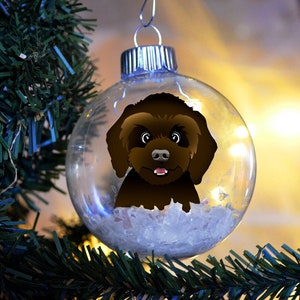 May include: A clear glass Christmas ornament with a brown dog illustration inside. The ornament is hanging on a green Christmas tree branch. The dog has a happy expression with a pink tongue.
