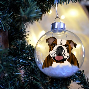 May include: Clear glass Christmas ornament featuring a brown and white bulldog illustration. The ornament hangs from a silver metal cap and is filled with white, fluffy material. The ornament is hanging on a green Christmas tree.