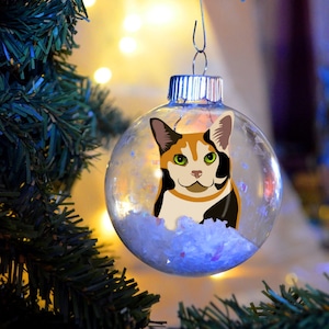 May include: Clear glass Christmas ornament with a calico cat design. The ornament is filled with white and blue decorative material and hangs from a silver metal hook. The ornament is hanging on a green Christmas tree.