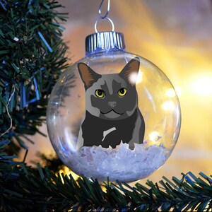 May include: A clear, round Christmas ornament featuring a gray cat illustration with yellow eyes. The ornament is suspended from a silver cap and filled with white, fluffy material. The ornament hangs in front of a blurred background with Christmas tree branches.