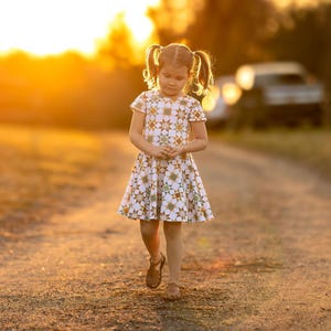 May include: A young child wearing a white dress with a yellow and green star pattern walks down a dirt path. The dress has short sleeves and a flared skirt. The child is wearing brown sandals. The background is blurred with a warm sunset.