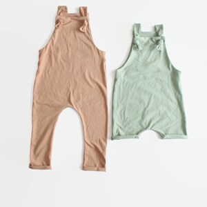 May include: Two sleeveless rompers, one in a muted tan color and one in a soft sage green. Both garments feature a high neckline, shoulder straps that tie into small knots at the front, and a relaxed fit with cuffed leg openings.