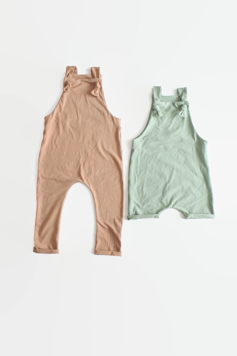 May include: Two sleeveless rompers, one in a muted tan color and one in a soft sage green. Both garments feature a high neckline, shoulder straps that tie into small knots at the front, and a relaxed fit with cuffed leg openings.