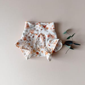 May include: Floral print baby bloomers with ruffled edges. The bloomers feature a white background with a pattern of orange, peach, and yellow flowers. A drawstring is visible at the waist. The bloomers are made of a soft fabric.