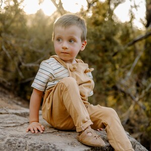 May include: A young child wearing tan overalls over a short-sleeved shirt with horizontal brown, blue, and white stripes. The child is also wearing tan leather sandals with a cutout design on the top, sitting on a textured rock surface.