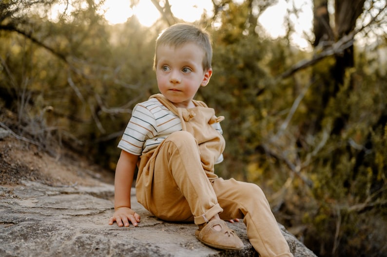 May include: A young child wearing tan overalls over a short-sleeved shirt with horizontal brown, blue, and white stripes. The child is also wearing tan leather sandals with a cutout design on the top, sitting on a textured rock surface.