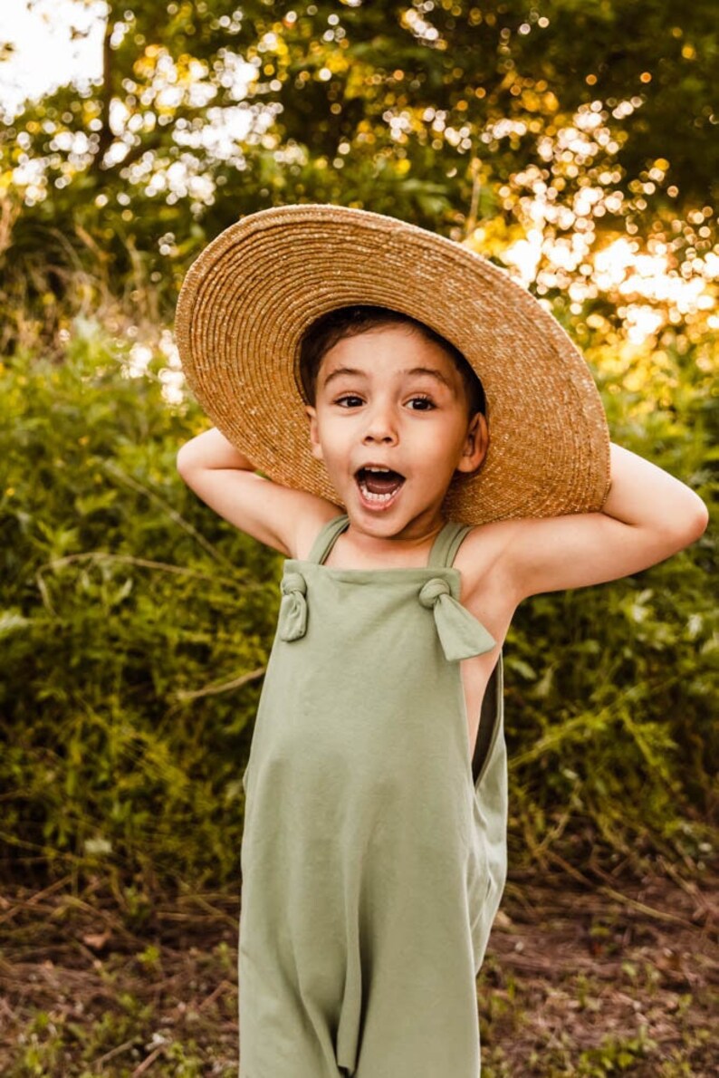 May include: A light olive green sleeveless romper featuring knotted shoulder straps, worn with a wide-brimmed natural straw hat. The romper has a simple, solid-colored design with a square neckline and a relaxed fit, paired with a textured straw hat.