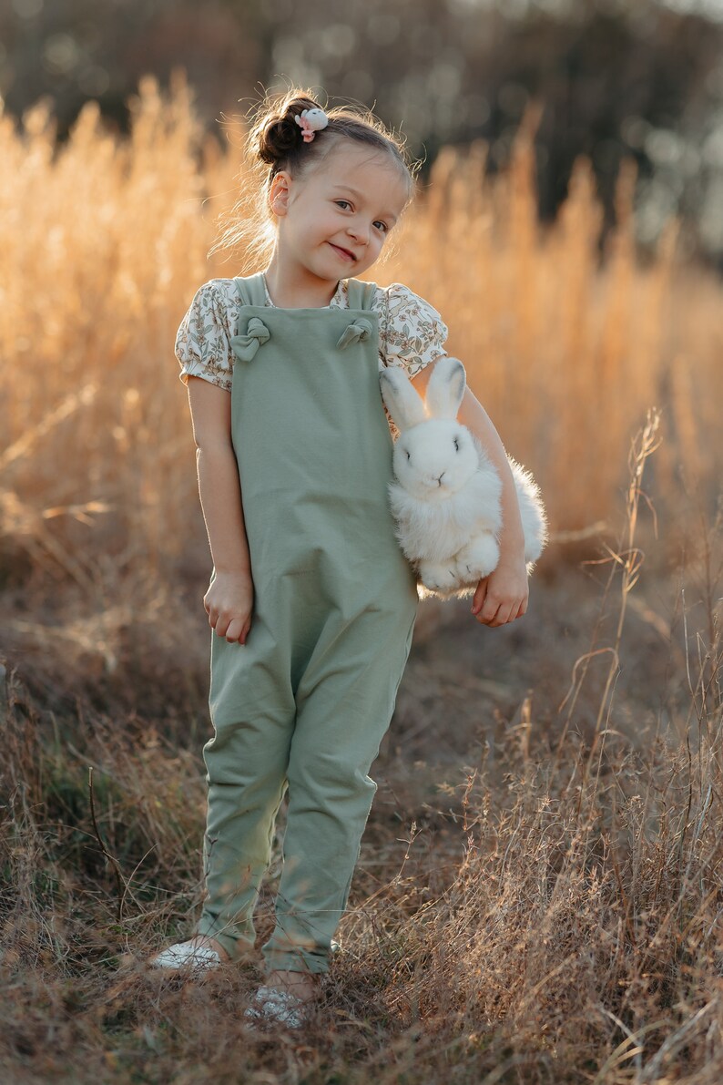 May include: A sage green jumpsuit with knotted shoulder straps worn over a short-sleeved shirt featuring a small floral pattern. The jumpsuit has a straight-leg design and is paired with a fluffy white plush rabbit toy held in one arm.