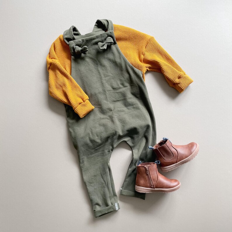May include: An olive green cotton overall with knotted shoulder straps layered over a mustard yellow long-sleeved waffle knit shirt, paired with a pair of brown leather ankle boots featuring side zippers and textured rubber soles.