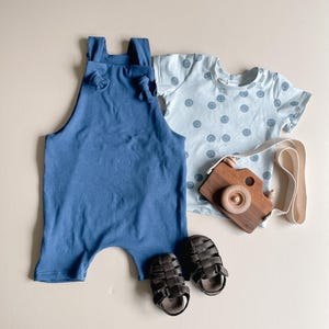 May include: A solid blue romper with knotted shoulder straps, a light blue short-sleeved t-shirt featuring a dark blue smiley face pattern, a pair of dark brown strappy sandals, and a wooden toy camera with a cream-colored fabric strap.