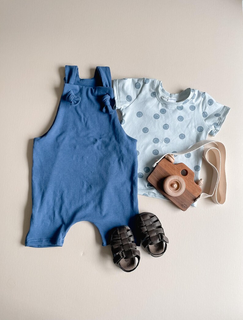 May include: A solid blue romper with knotted shoulder straps, a light blue short-sleeved t-shirt featuring a dark blue smiley face pattern, a pair of dark brown strappy sandals, and a wooden toy camera with a cream-colored fabric strap.