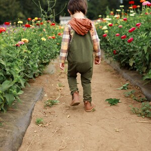May include: A child wearing olive green overalls over a long-sleeved plaid shirt with a rust-colored hood, paired with brown leather boots. The child is walking away from the camera on a dirt path between rows of colorful zinnia flowers.